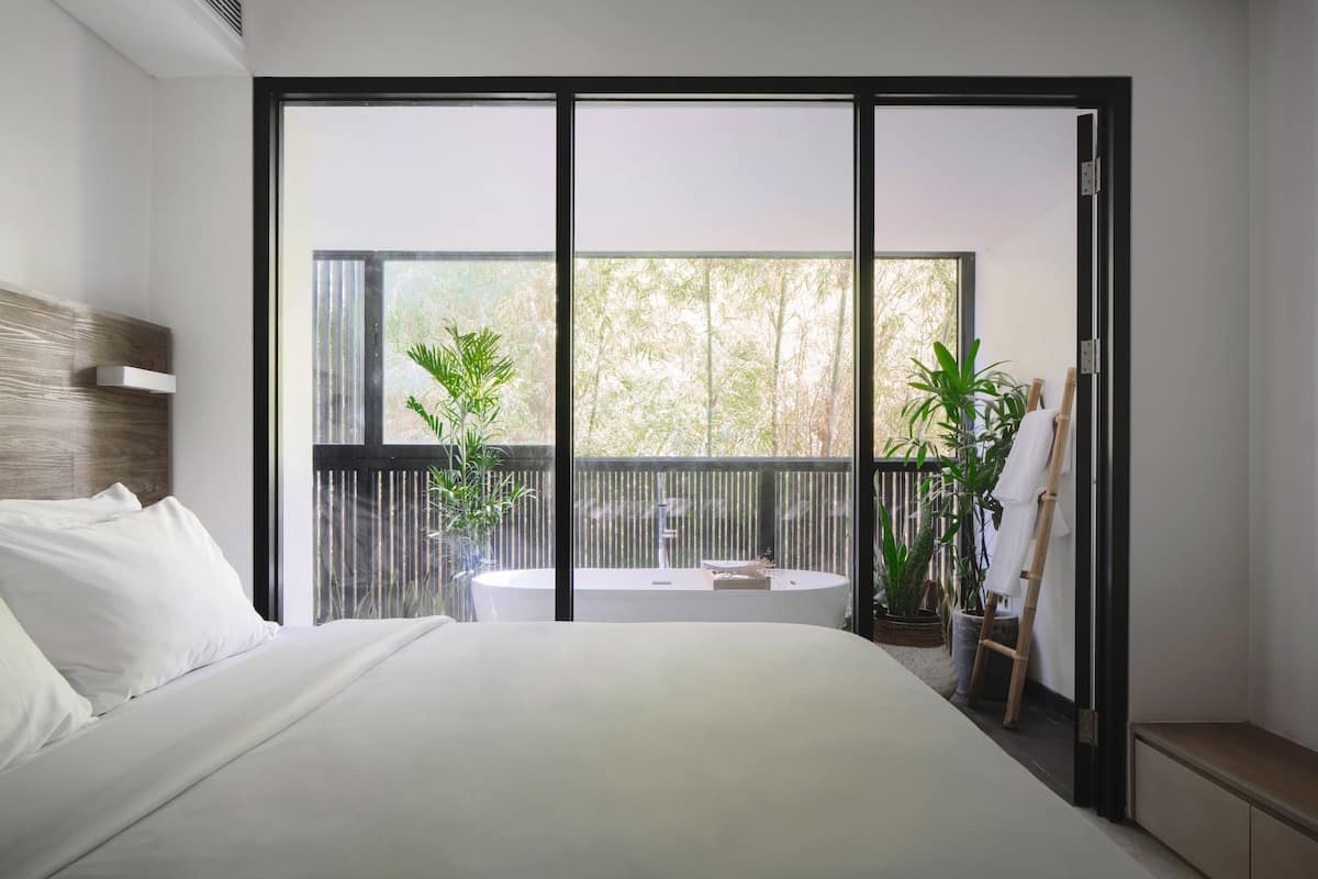 A modern bedroom with a large bed, a view of a balcony with plants, and a bathtub.