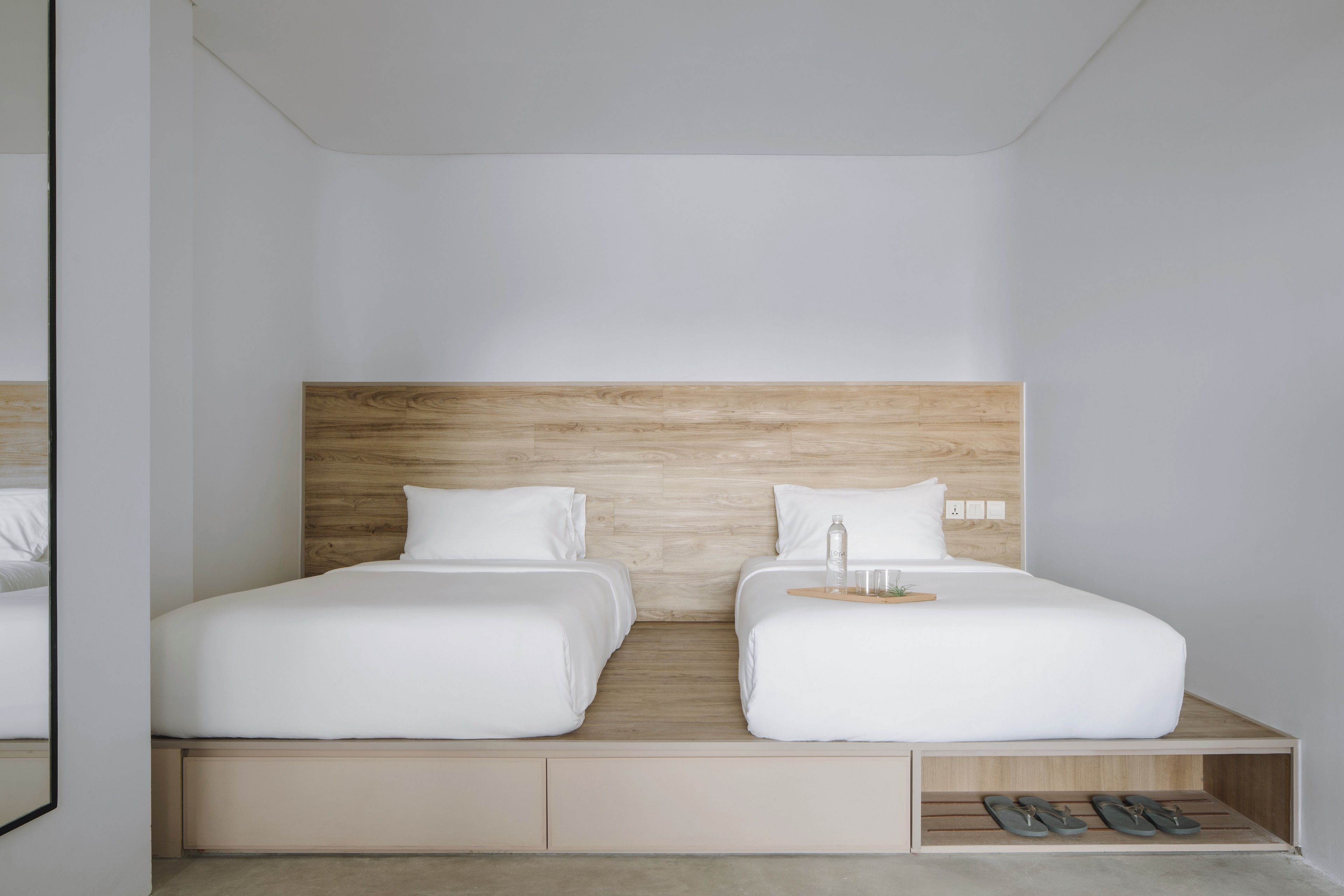 Una habitación de hotel moderna con dos camas individuales, un cabecero de madera, un pequeño estante con zapatillas y una mesita de noche con una botella y vasos.