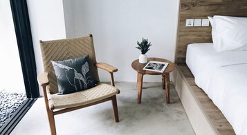 Una stanza con una sedia di legno, un tavolino rotondo con una pianta e un letto con lenzuola bianche.