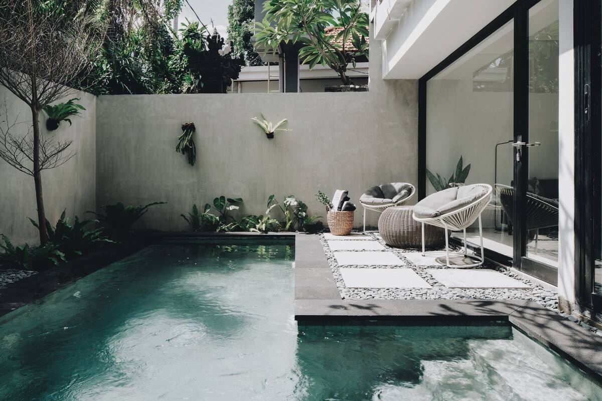 A modern outdoor pool area with a glass enclosure, surrounded by plants and furnished with wicker chairs and a wicker basket.