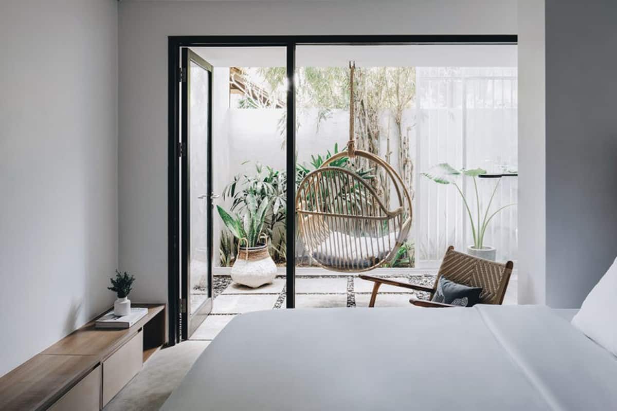 A modern bedroom with a large bed, a wooden headboard, a wicker chair, and a view of a garden through a sliding glass door.