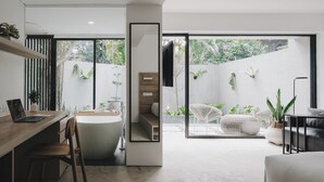 A modern bathroom with a bathtub, a wooden desk, and a chair. There is a large window with a view of greenery outside. - Lloyd's Inn Bali (Seminyak)