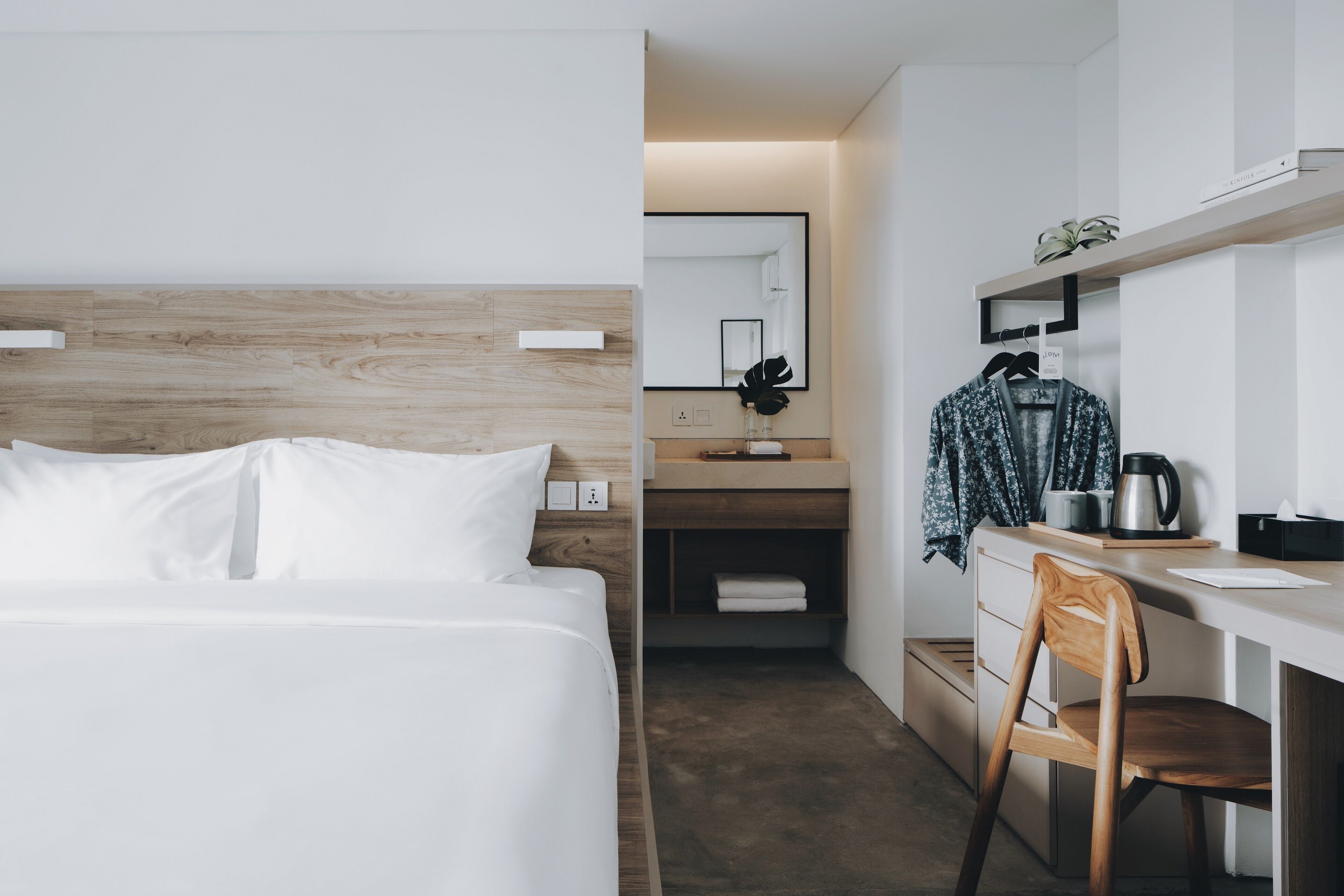 A modern hotel room with a wooden headboard, a white bed, a wooden chair, a small kitchenette, and a mirror on the wall.