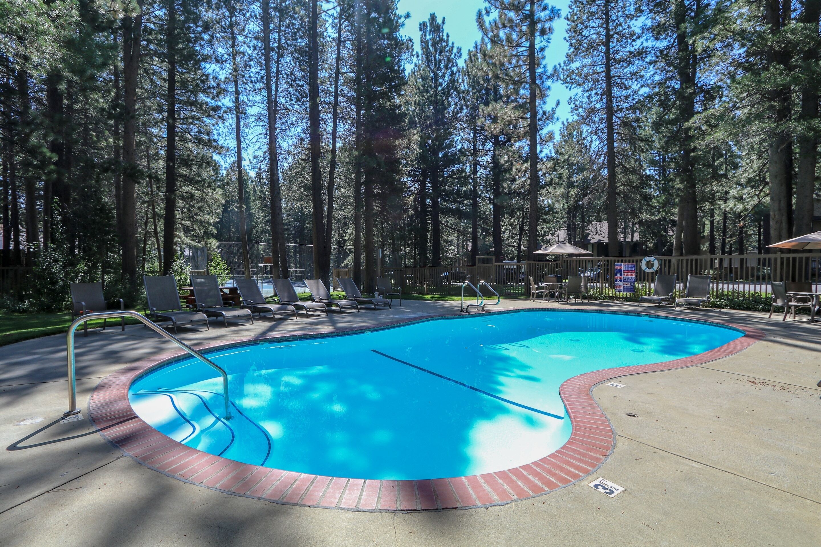 A swimming pool with a brick edge, surrounded by lounge chairs and a forested area.
