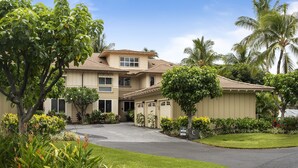A two-story building with a beige exterior, a curved driveway, and lush landscaping including palm trees and flowering plants. - Waikoloa Beach S C3 2 Bedroom Villa by RedAwning (Waikoloa)