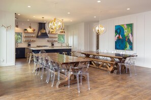 A spacious dining area with a large wooden table and clear plastic chairs, a kitchen with white cabinets and a black range hood, and a colorful abstract painting on the wall. - The White House On The Horseshoe 8 Bedroom Home by RedAwning (Canyon Lake)