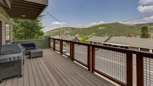 Een houten terras met een barbecue en tuinmeubilair, met uitzicht op een woonwijk met huizen en heuvels.