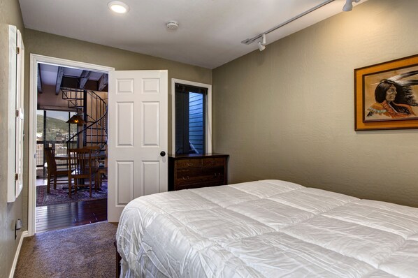A bedroom with a white bed, a wooden dresser, a door leading to a balcony, and a painting on the wall.