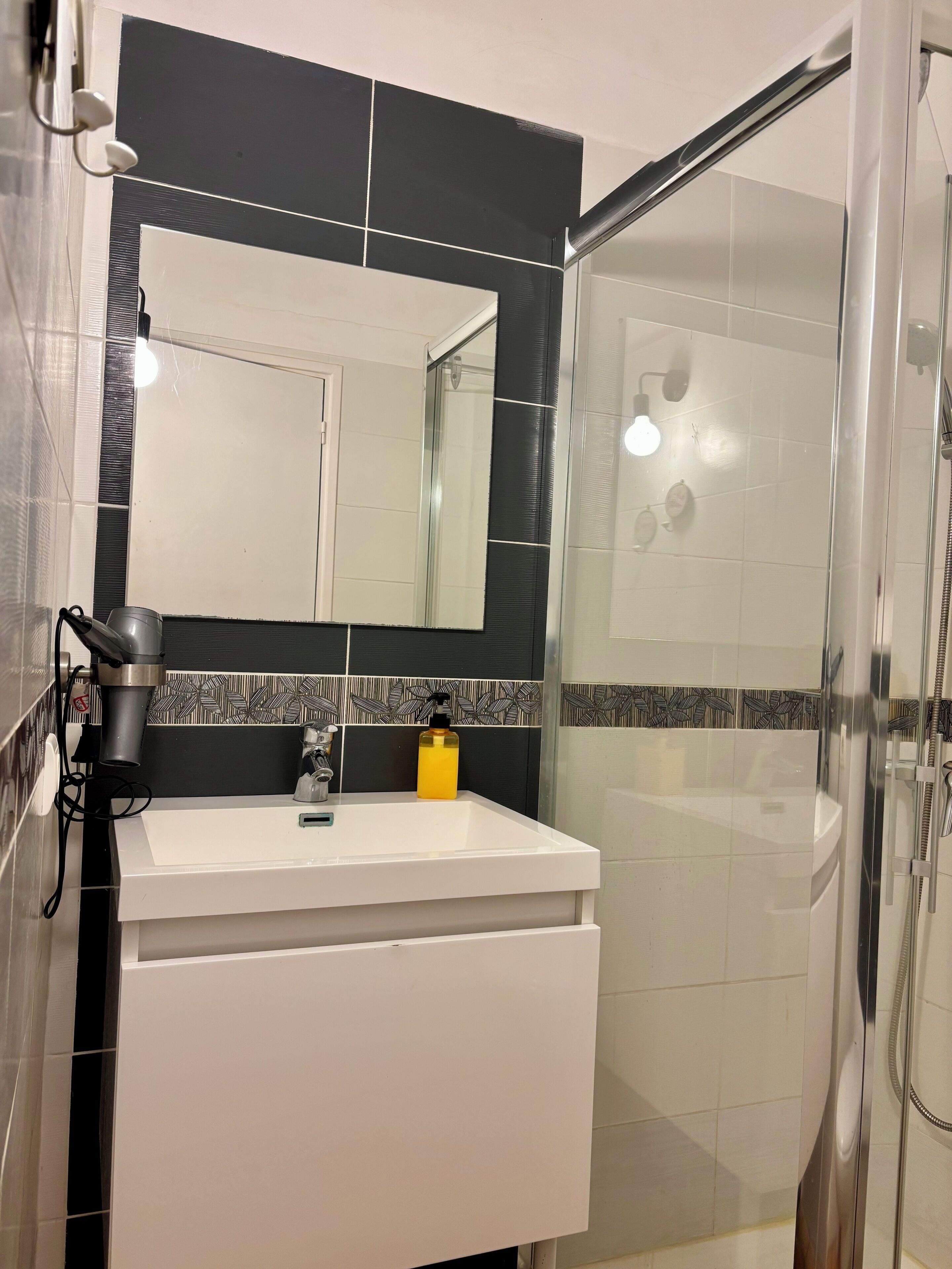 Een badkamer met een wit wastafelmeubel, een spiegel, een douchecabine en een betegelde spatwand met een patroon.