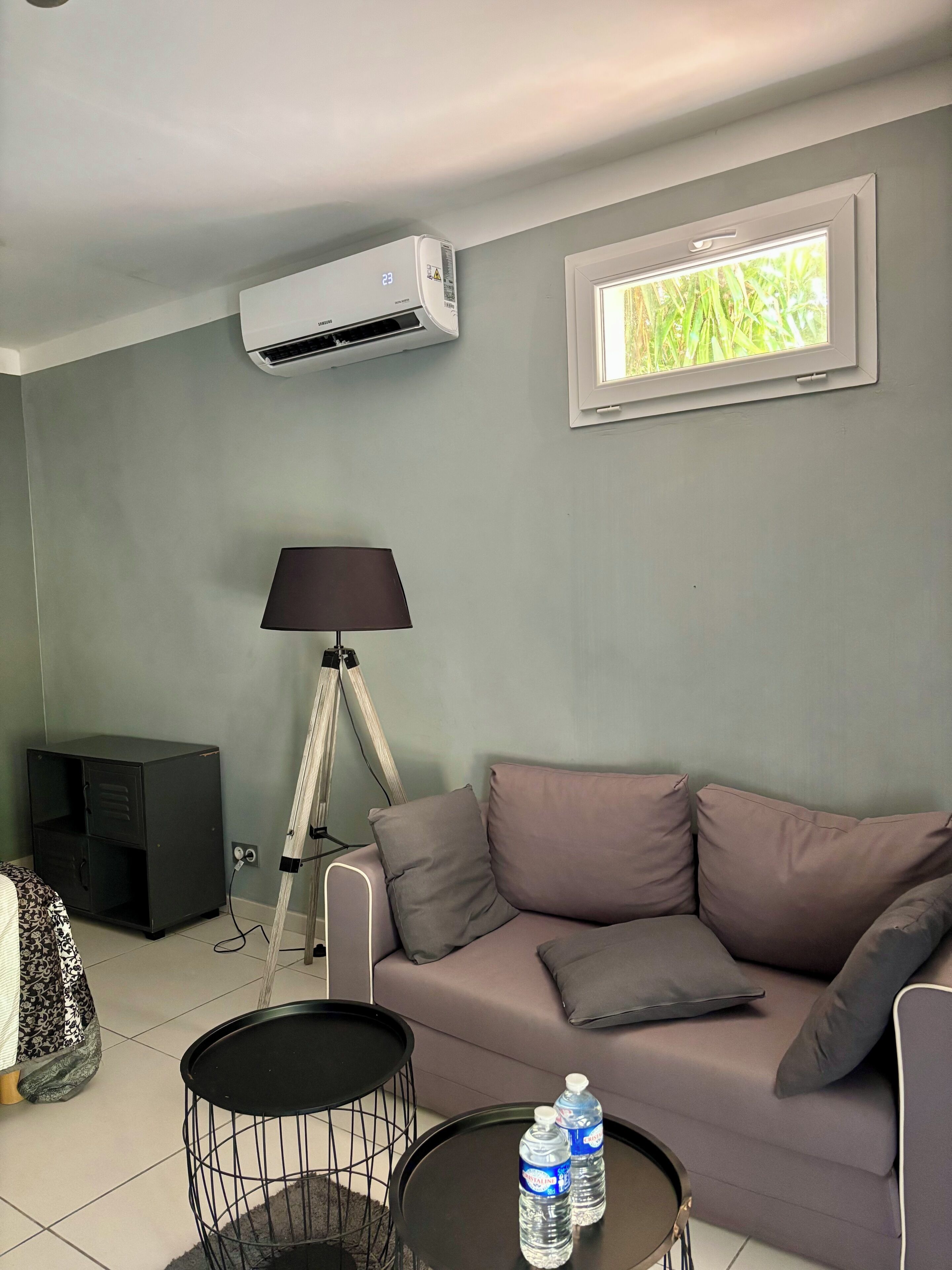 A living room with a grey sofa, a small round coffee table, a lamp, an air conditioning unit, and a window with a view of greenery.