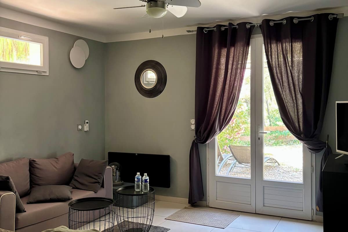 Une pièce dotée d’un ventilateur de plafond, d’un canapé, d’une table basse, d’un téléviseur, d’une fenêtre avec des rideaux et d’une porte donnant sur l’extérieur.
