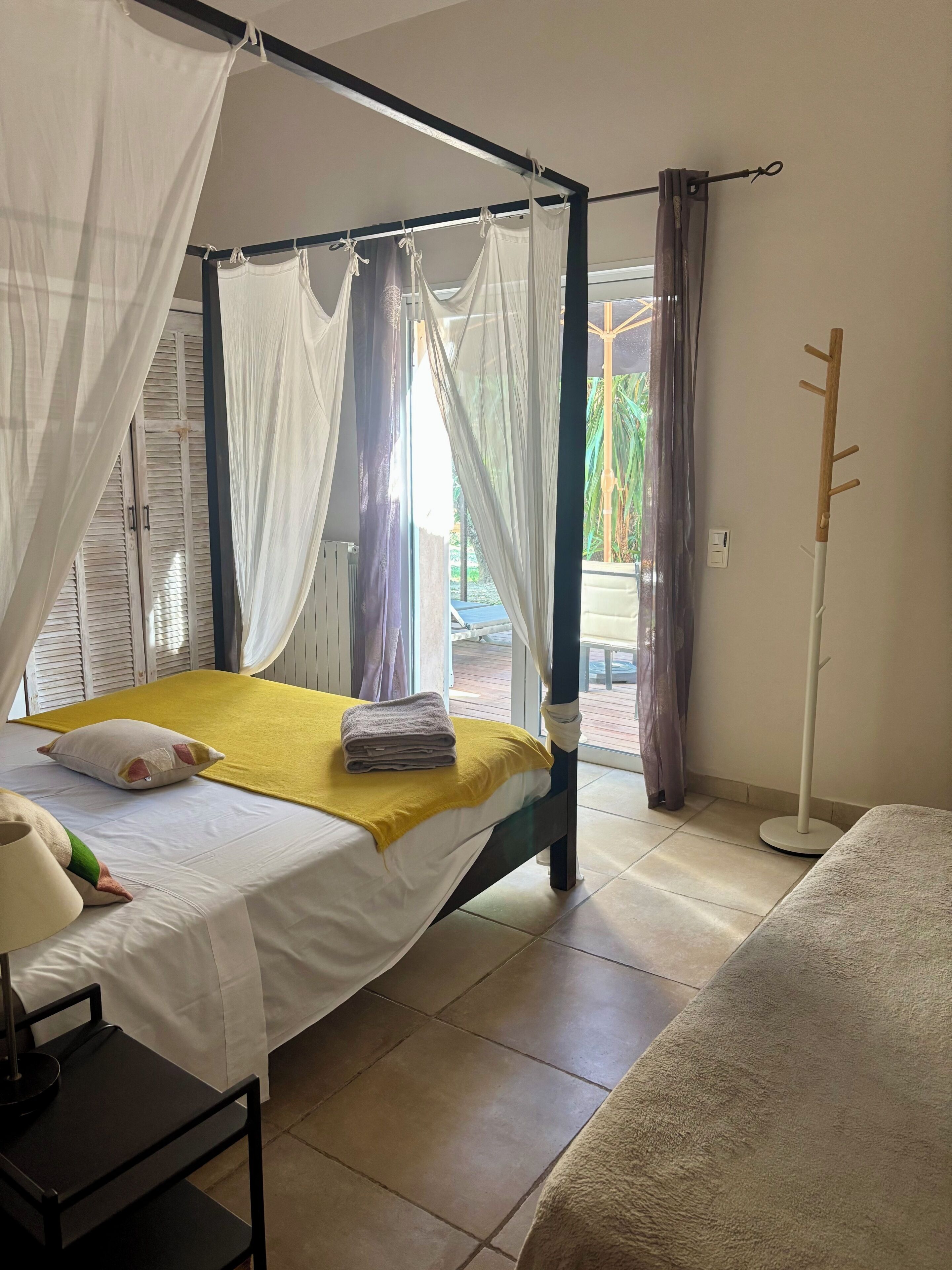 A room with a bed, a bedside table, a floor lamp, and a view of the outside.