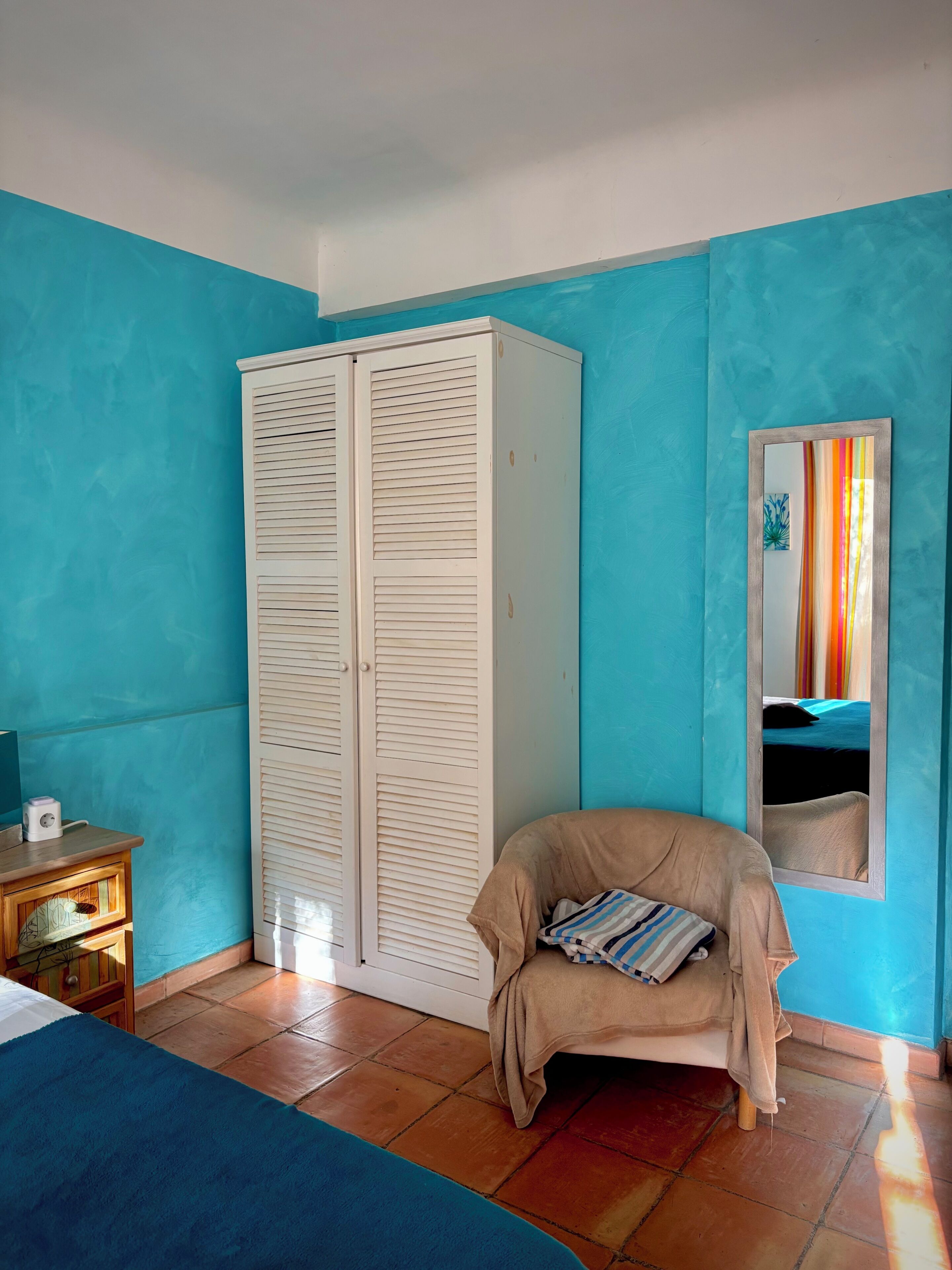 Una habitación con pared color turquesa, un armario blanco, una cama con cubrecolor beige y una mesita de noche de madera con una lámpara.