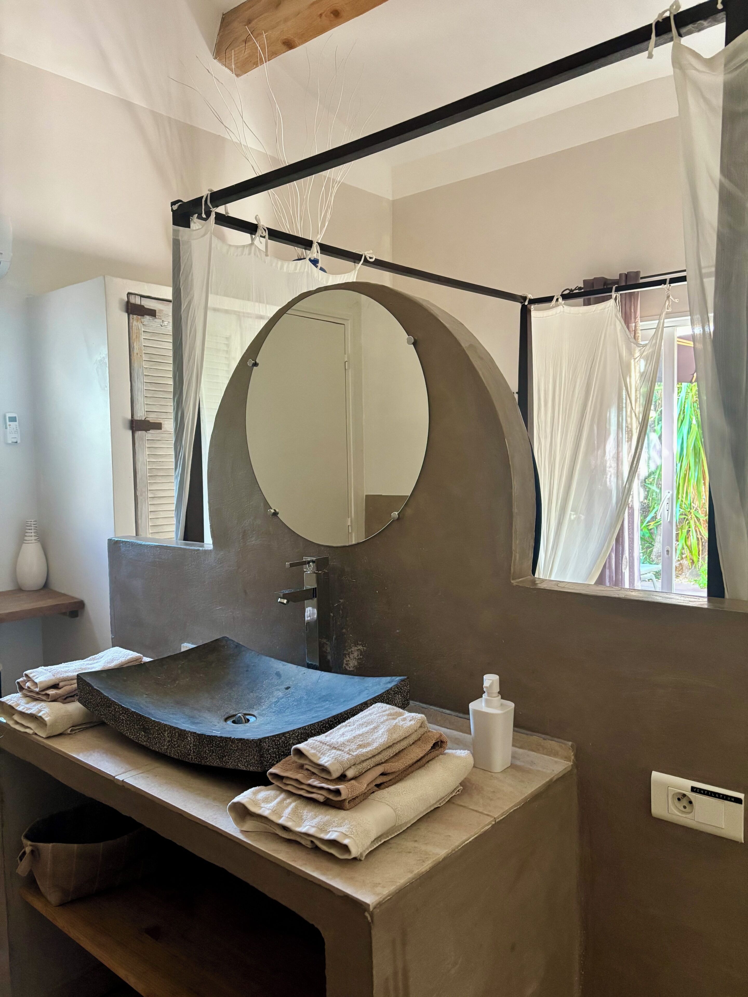 A bathroom with a round mirror, a sink, towels, a bottle, and a window with curtains.