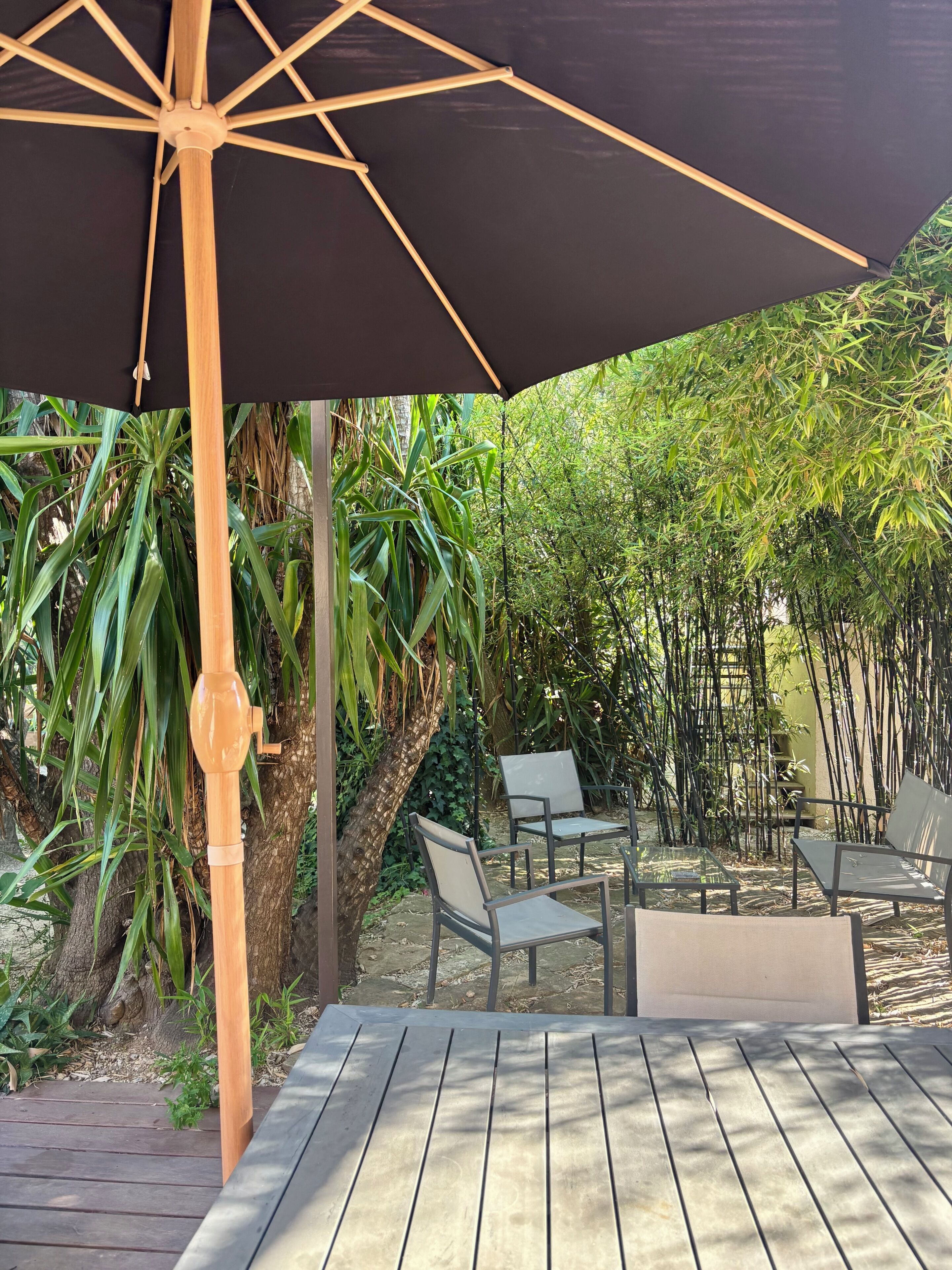 An outdoor seating area with a wooden table and chairs, a large umbrella, and lush greenery in the background.