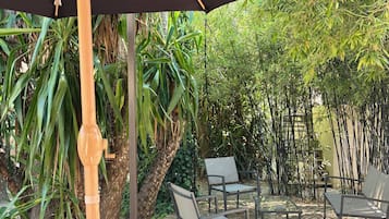 An outdoor seating area with a wooden table and chairs, a large umbrella, and lush greenery in the background.