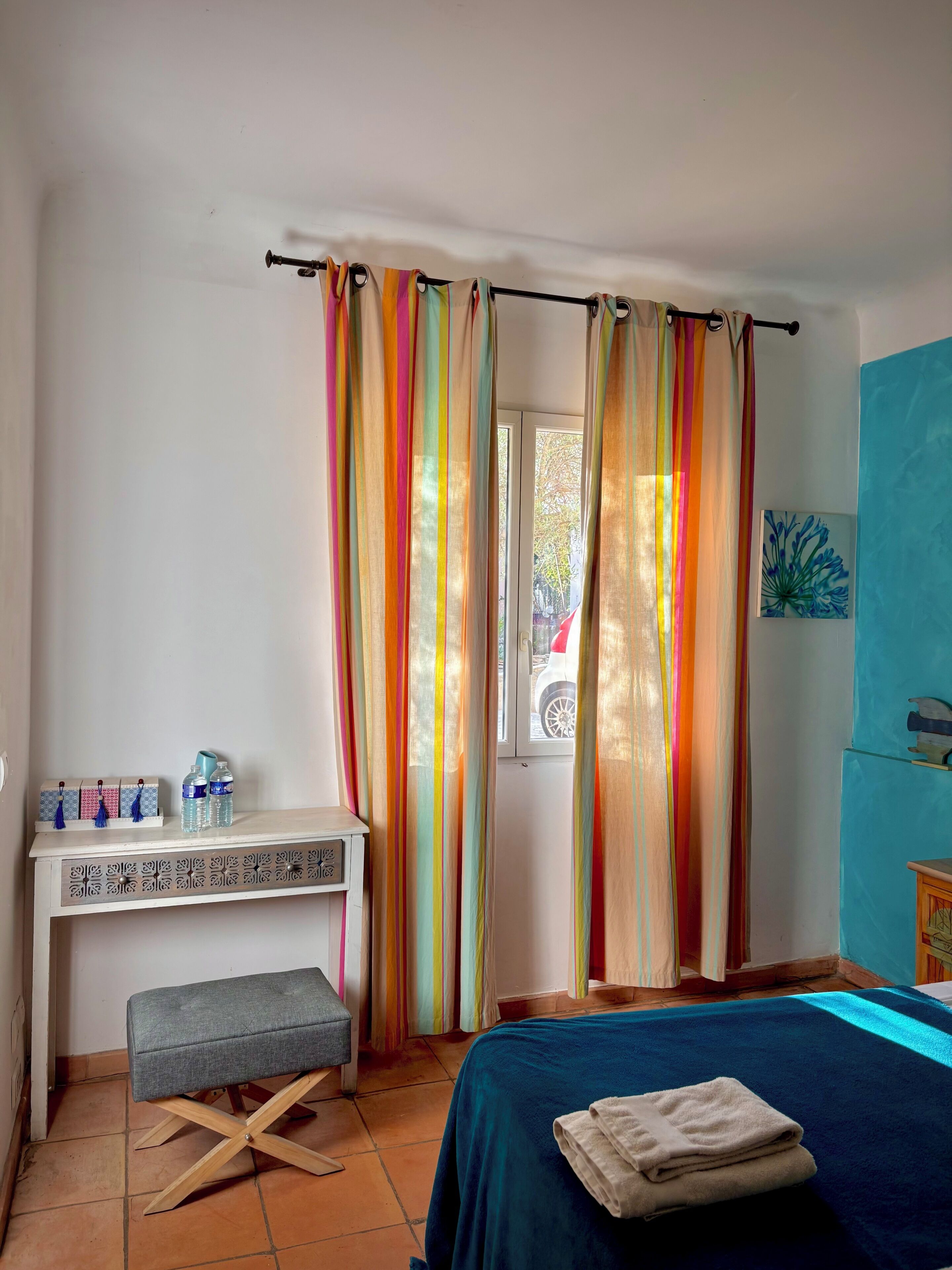 A room with a bed, a desk with a bench, a window with curtains, and a wall with a picture.
