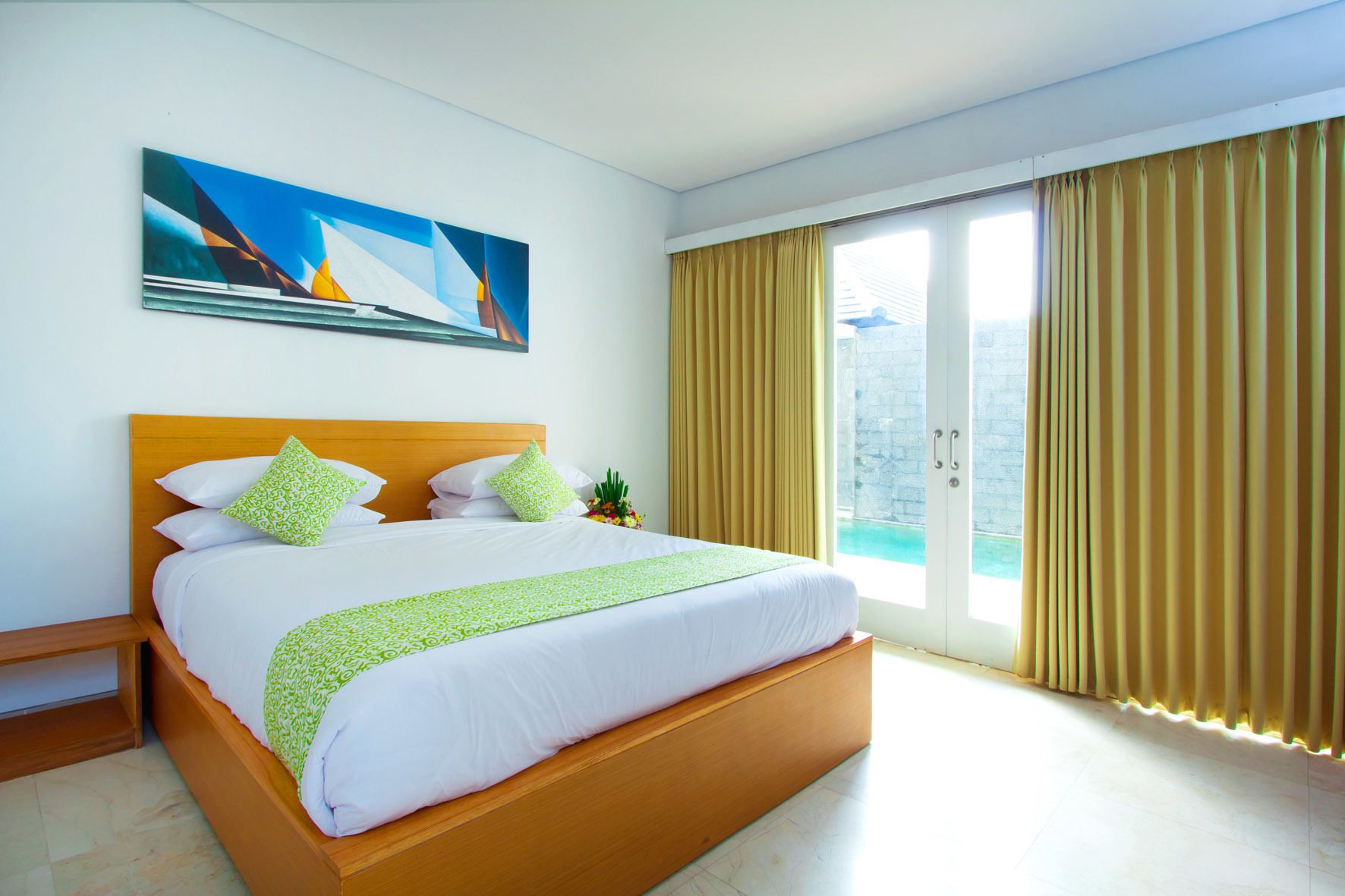 A hotel room with a large bed, a wooden headboard, a painting on the wall, and a view of a pool through the window.