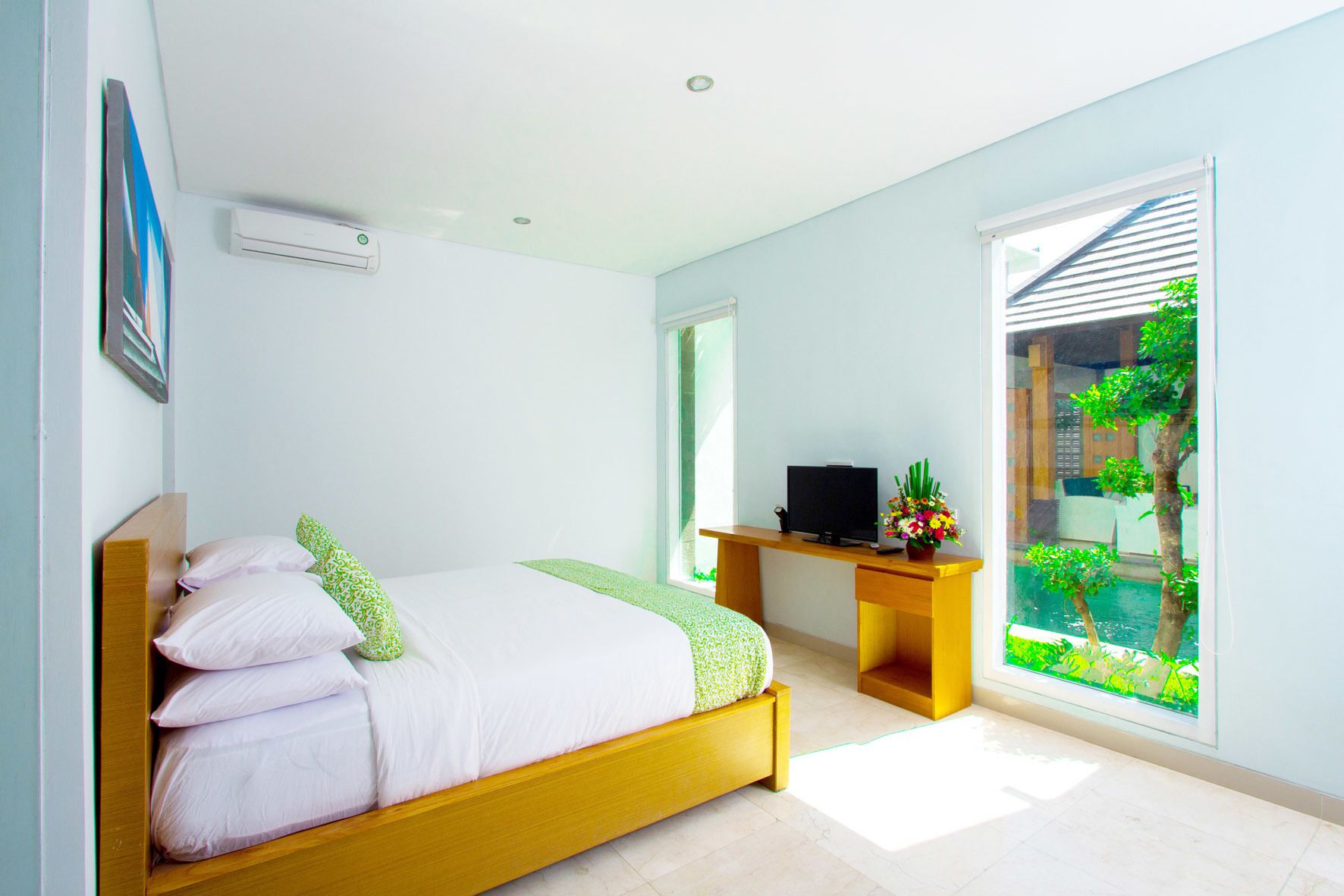 A modern bedroom with a bed, a desk, and a window with a view of greenery and buildings.