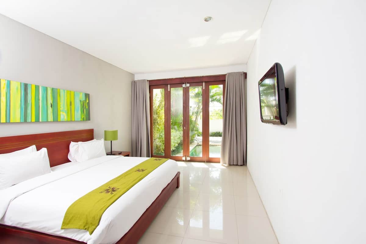A hotel room with a bed, a TV, a painting, and a view of greenery outside.
