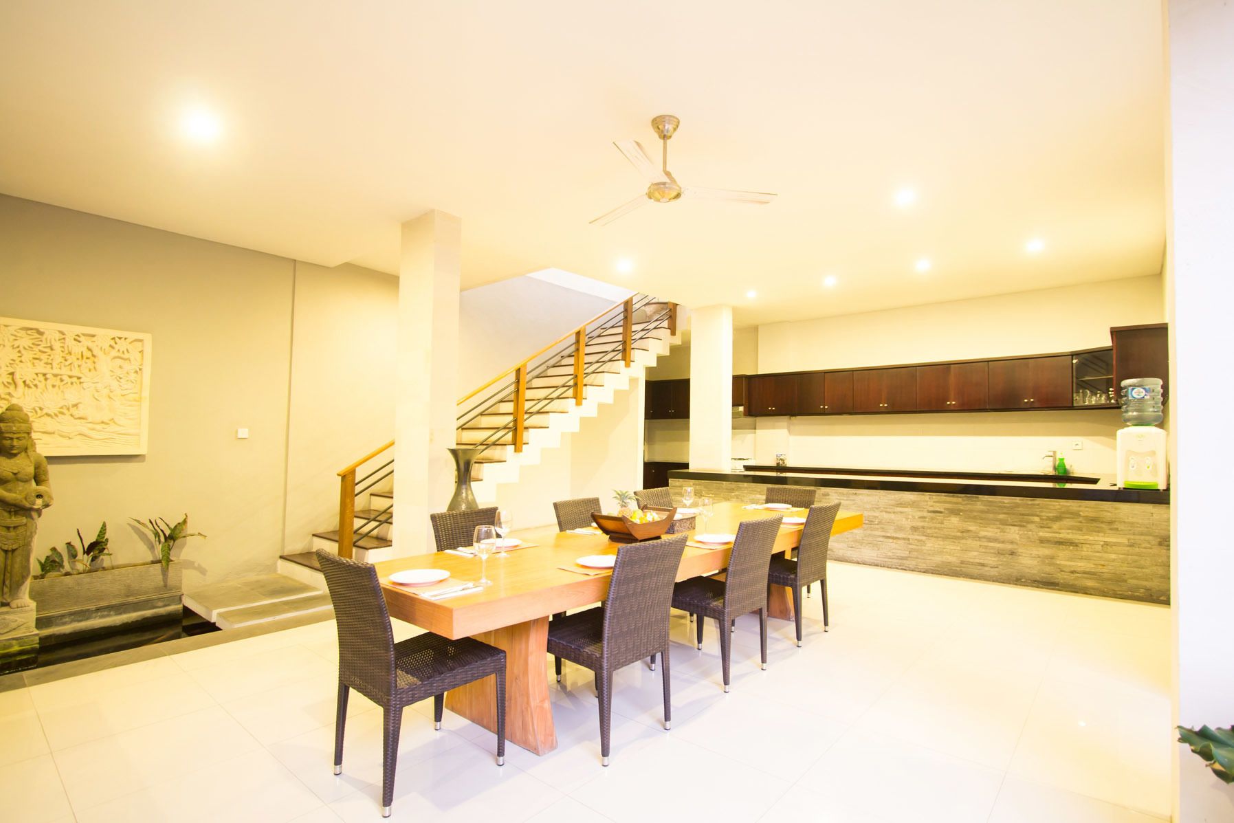 A modern dining area with a wooden table and wicker chairs, a staircase, and a kitchen in the background.