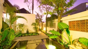 A serene garden with a water feature, lush greenery, and a pathway leading to a building with a decorative wall. - Apple Villa and Apartment (Kerobokan)