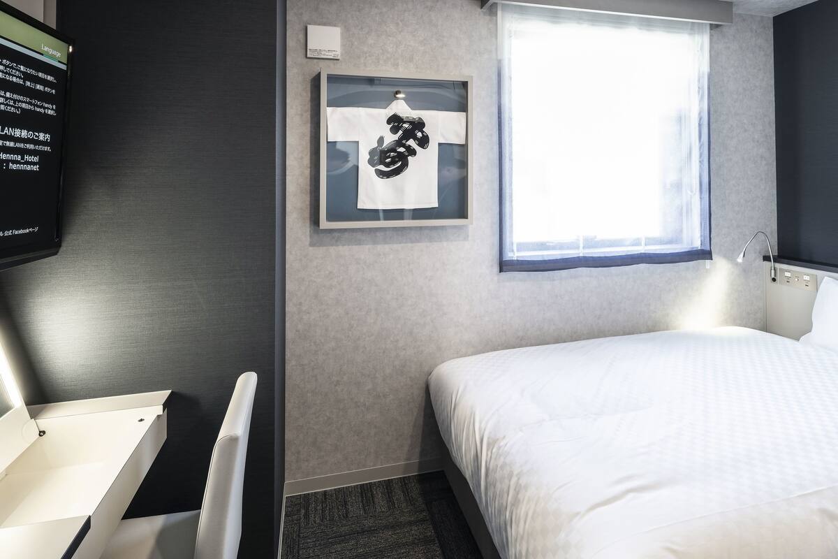Ein Hotelzimmer mit Bett, Schreibtisch, Stuhl, einem gerahmten Bild an der Wand und einem fenster mit Vorhängen.