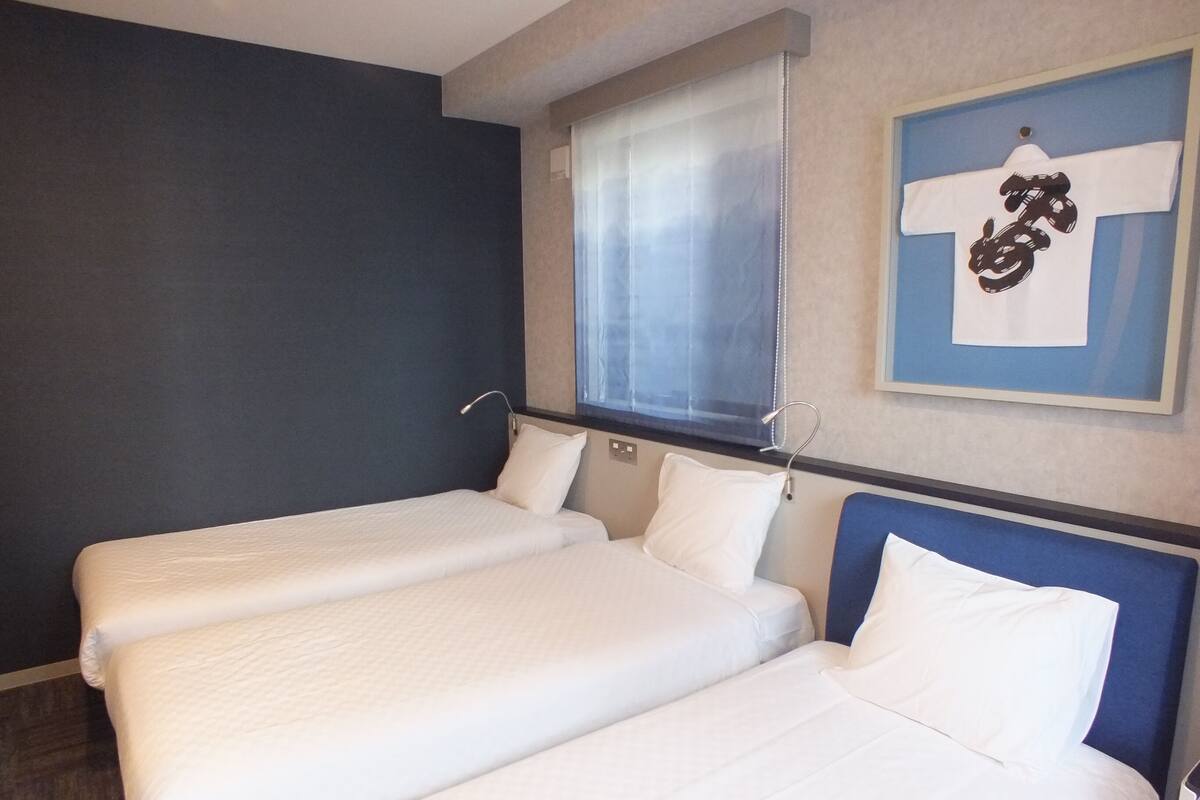 Ein Hotelzimmer mit zwei Einzelbetten, einem Fenster mit Jalousien, einem Wandbild und einem blauen Kopfteil.