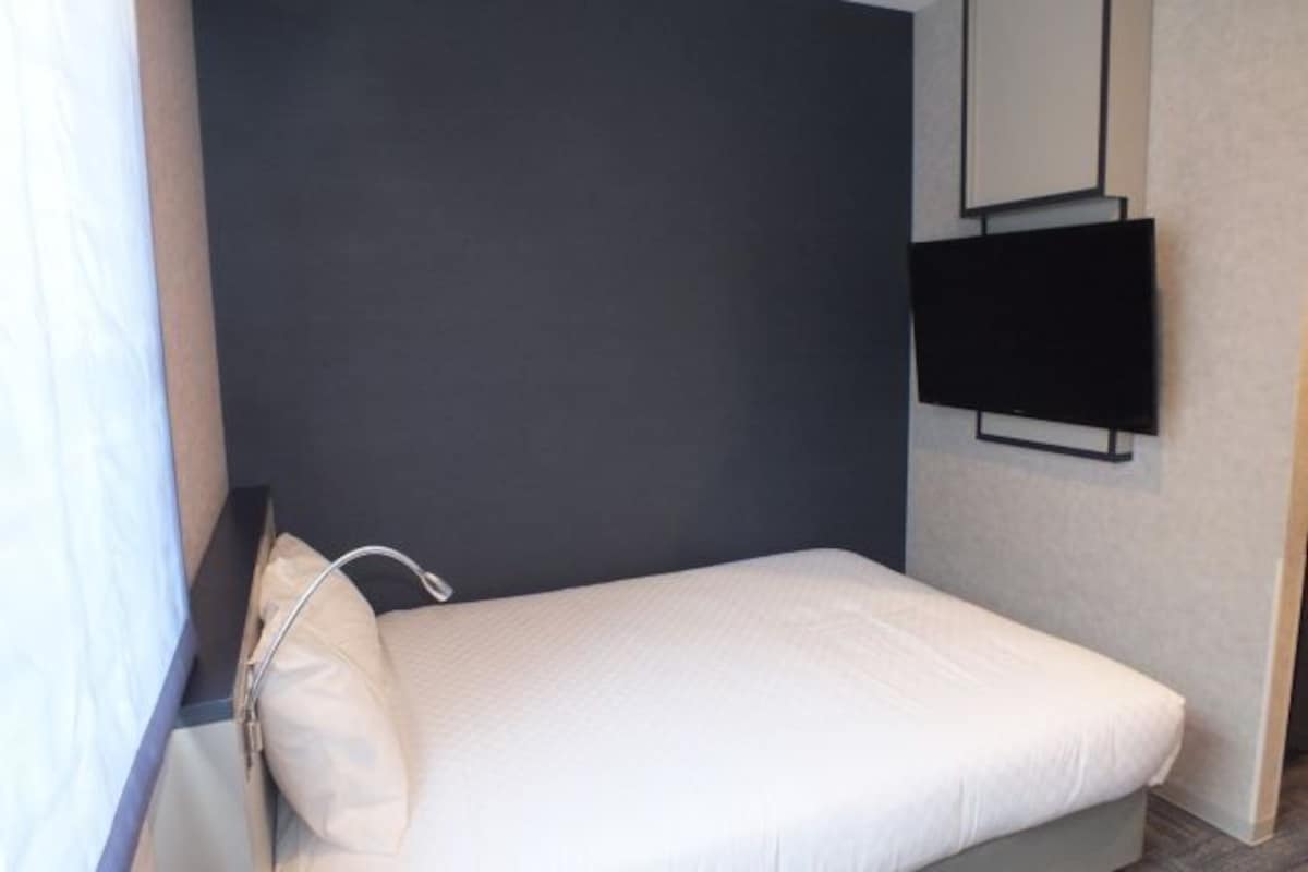 Una habitación pequeña con cama individual, pared oscura, televisor montado y ventana con cortina.