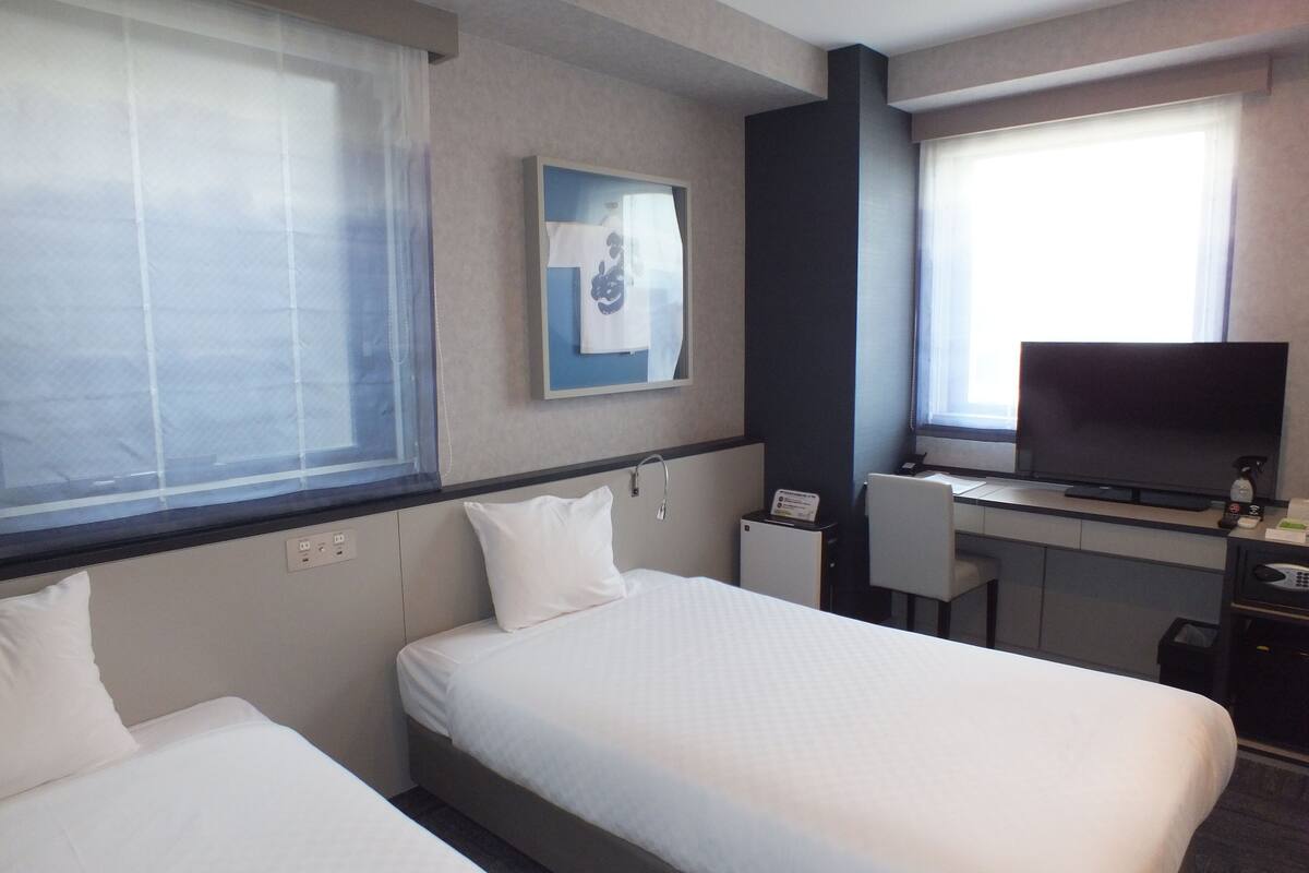 Ein Hotelzimmer mit zwei Betten, einem Schreibtisch mit Fernseher und einem Fenster mit Jalousien.