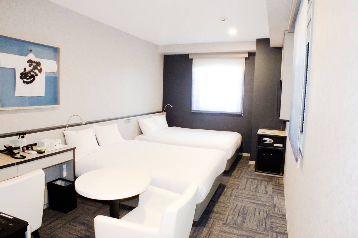 Ein Hotelzimmer mit Bett, einem kleinen Schreibtisch mit Stuhl, einem runden Tisch und einem Fenster mit Jalousien.