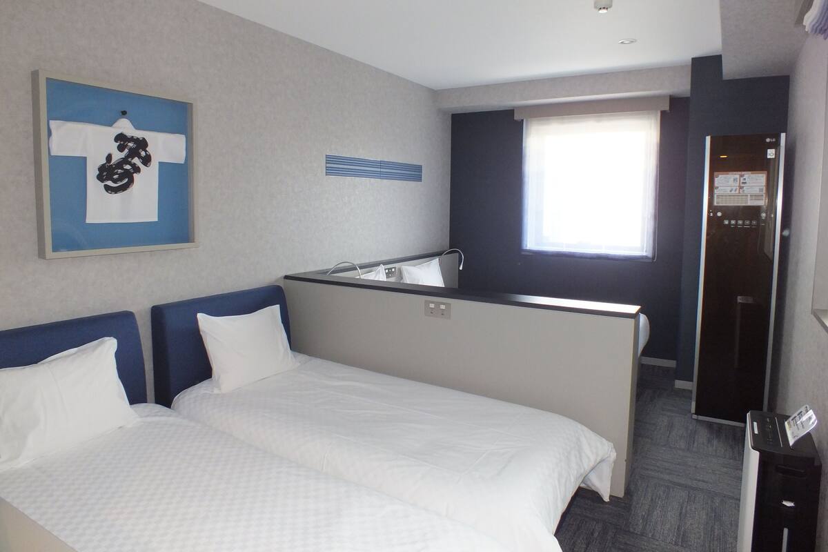 Ein Hotelzimmer mit zwei Betten, einer kleinen Küchenzeile, einem Fenster mit Jalousien und einer Tür mit Gegensprechanlage.