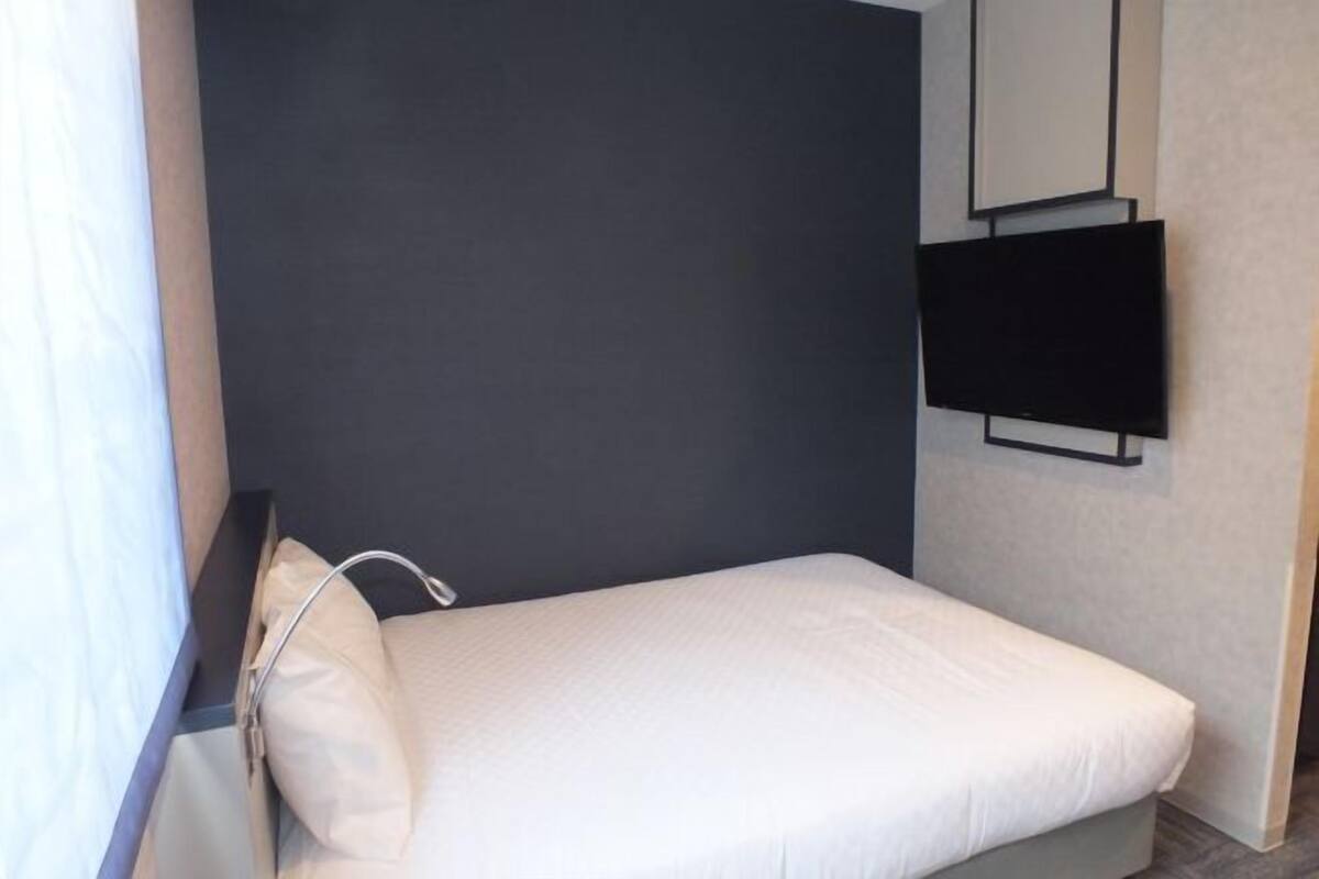 Ein kleines Zimmer mit einem Einzelbett, einer dunklen Wand, einem an der Wand befestigten Fernseher und einem Fenster mit Vorhang.