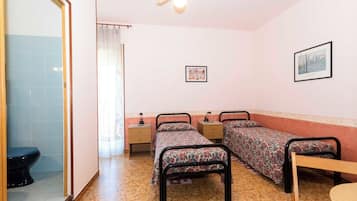 Un dormitorio con dos camas, una mesa de madera, un ventilador de techo, un cuadro enmarcado en la pared y un baño con inodoro y lavabo.