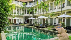 A pool area with lounge chairs and umbrellas, surrounded by greenery and a multi-story building with balconies. - WORLD WALKERs - Hostel (Siem Reap)