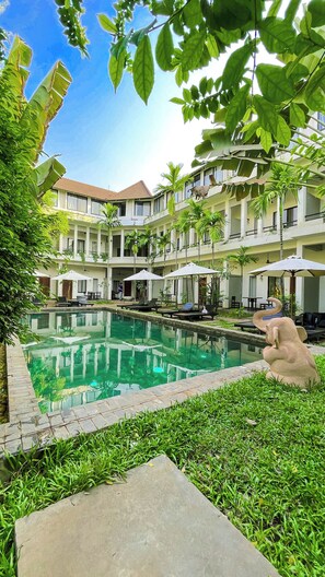 A pool area with lounge chairs and umbrellas, surrounded by greenery and a multi-story building with balconies. - WORLD WALKERs - Hostel (Siem Reap)
