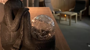 A statue of a seated figure with a textured robe, placed on a wooden table. In the background, there is a room with a wooden wall, a round mirror, and two chairs.