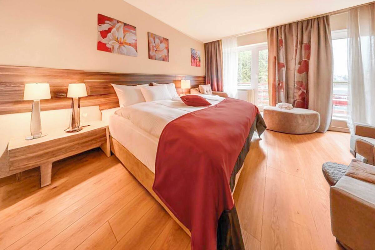 Une chambre d’hôtel avec une tête de lit en bois, un grand lit recouvert d’une couverture rouge, deux tables de chevet avec des lampes, un petit pouf rond et une fenêtre avec des rideaux.