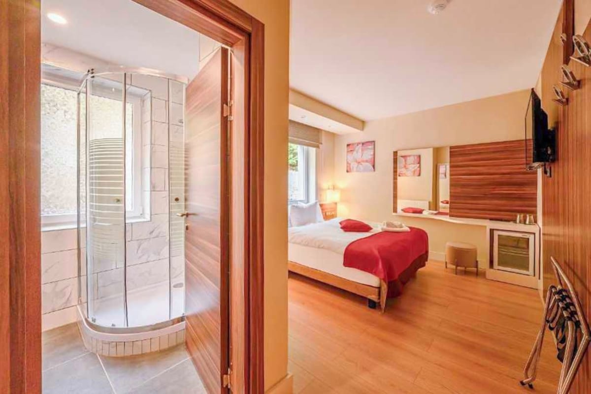 A hotel room with a glass-enclosed shower, a bed with a red blanket, a wooden cabinet, and a large mirror.
