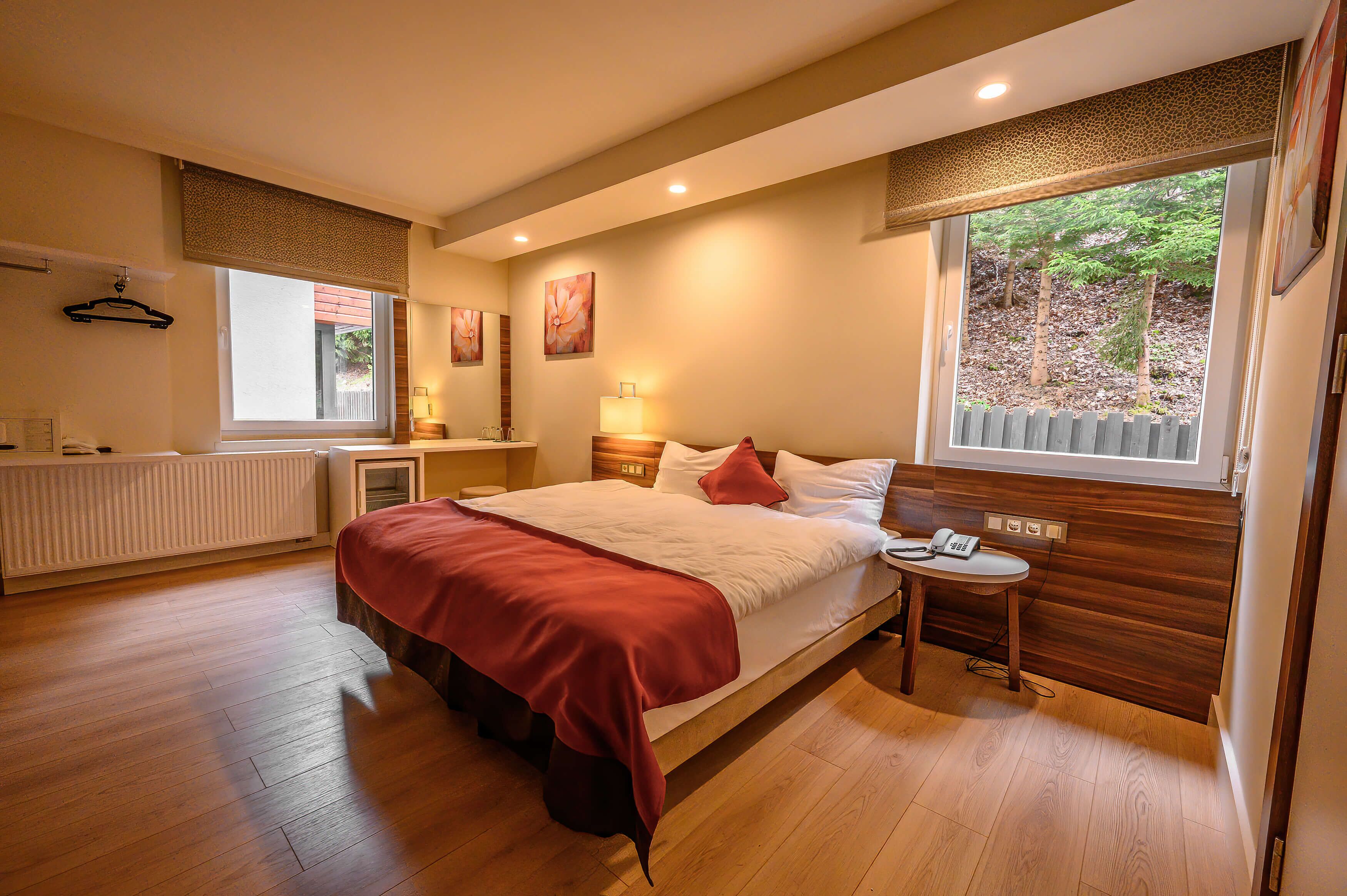Een slaapkamer met een groot bed, een nachtkastje met een lamp, een radiator en een raam met uitzicht op bomen.