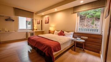 Een slaapkamer met een groot bed, een nachtkastje met een lamp, een radiator en een raam met uitzicht op bomen.
