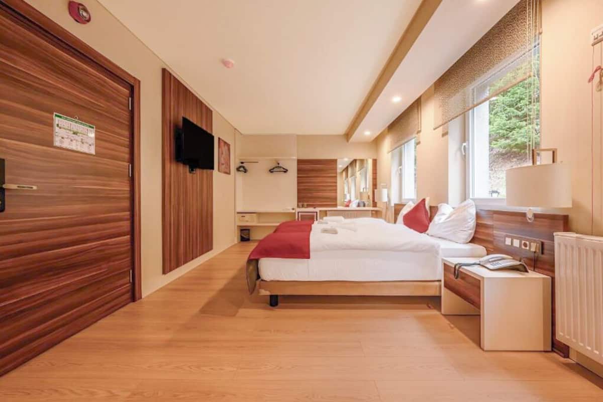 Une chambre d’hôtel moderne avec un plancher en bois, un lit recouvert de linge de maison blanc, une table de chevet avec une lampe et une fenêtre donnant sur des arbres.