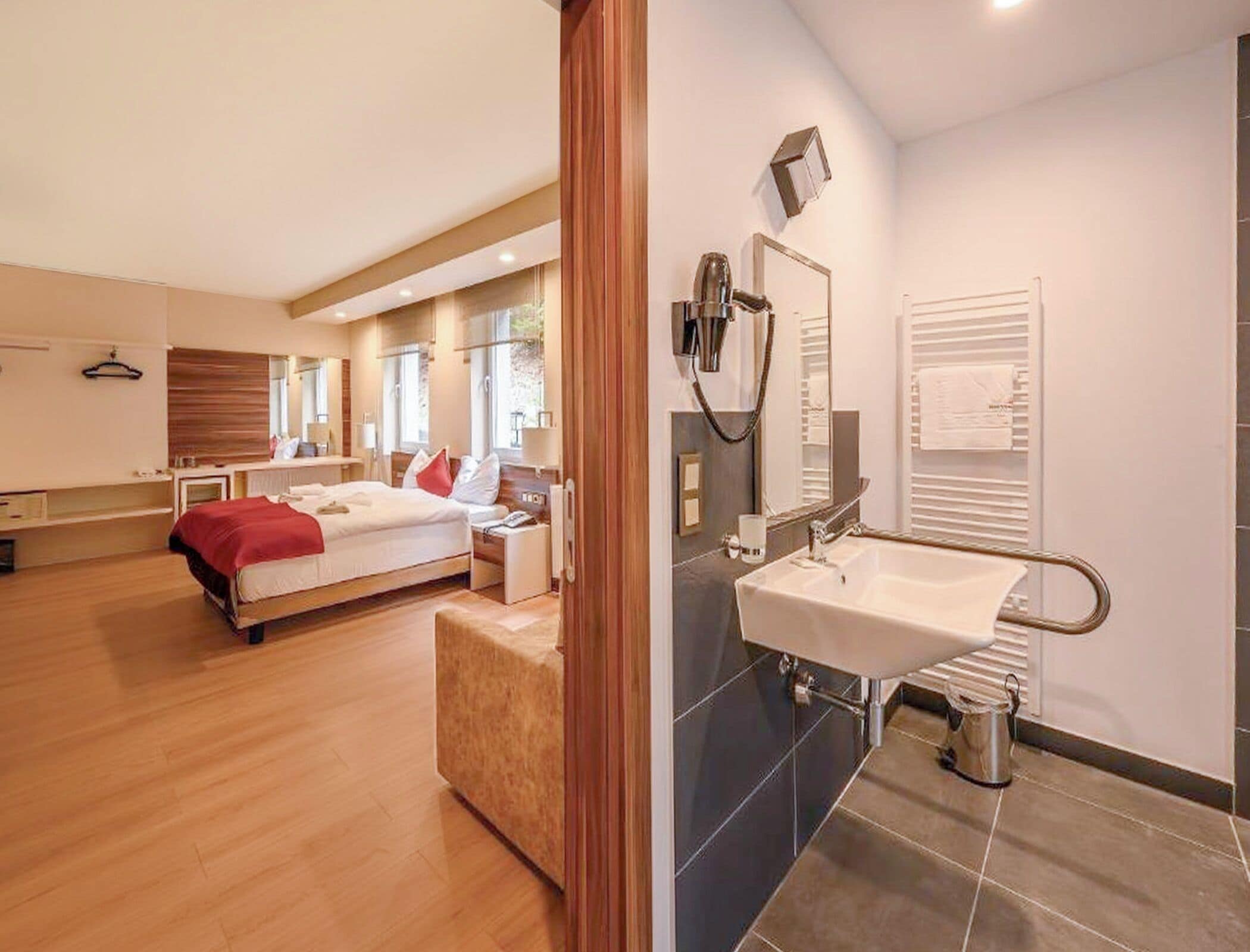 A modern hotel room with a large bed, a bedside table, a mirror, a bathroom with a sink and shower, and a wooden floor.