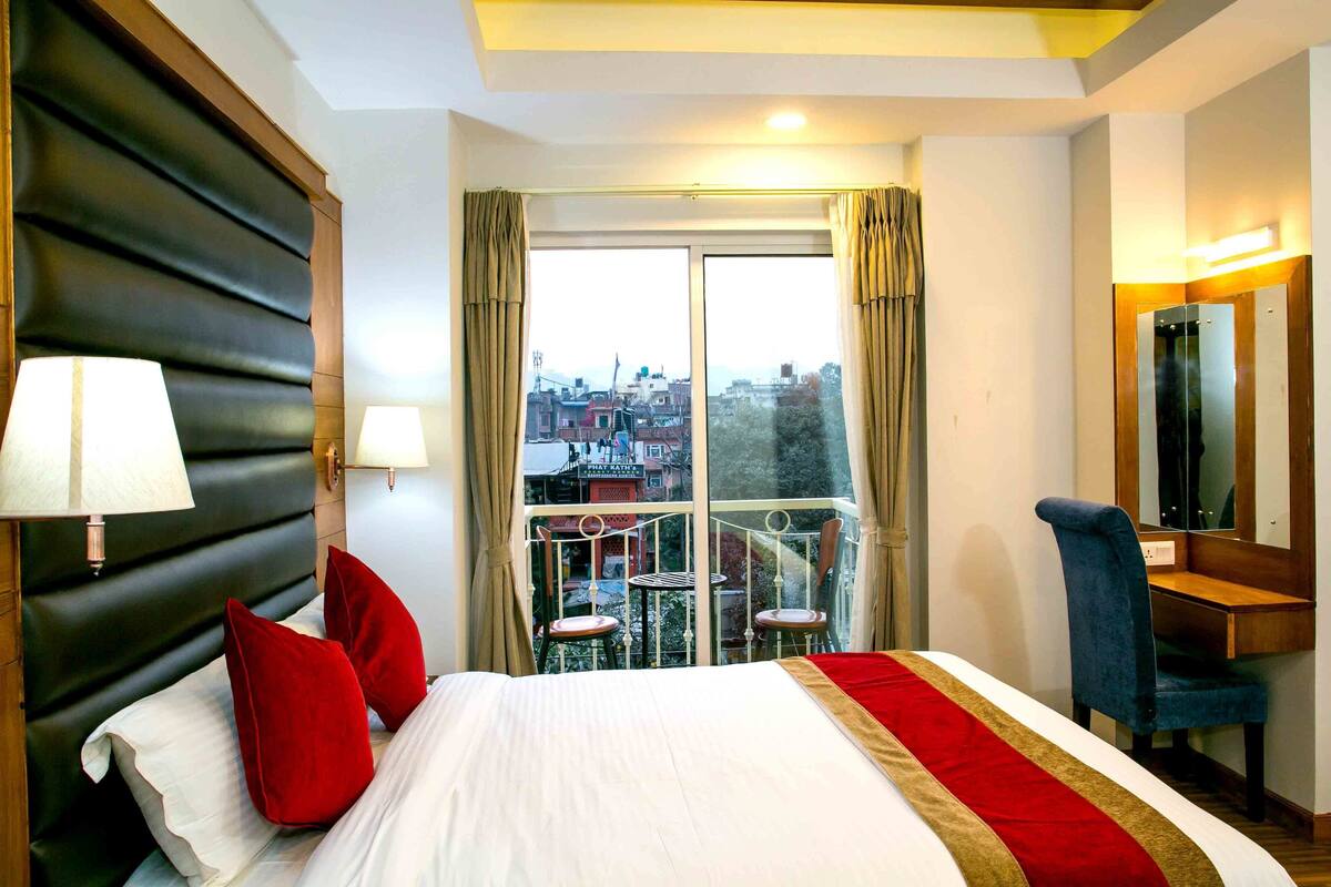 A hotel room with a large bed, a desk with a chair, a balcony with a view of the city, and a large mirror.