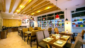 A restaurant with wooden ceiling beams, multiple dining tables with chairs, and a kitchen area in the background. - Beautiful Kathmandu Hotel (Kathmandu)