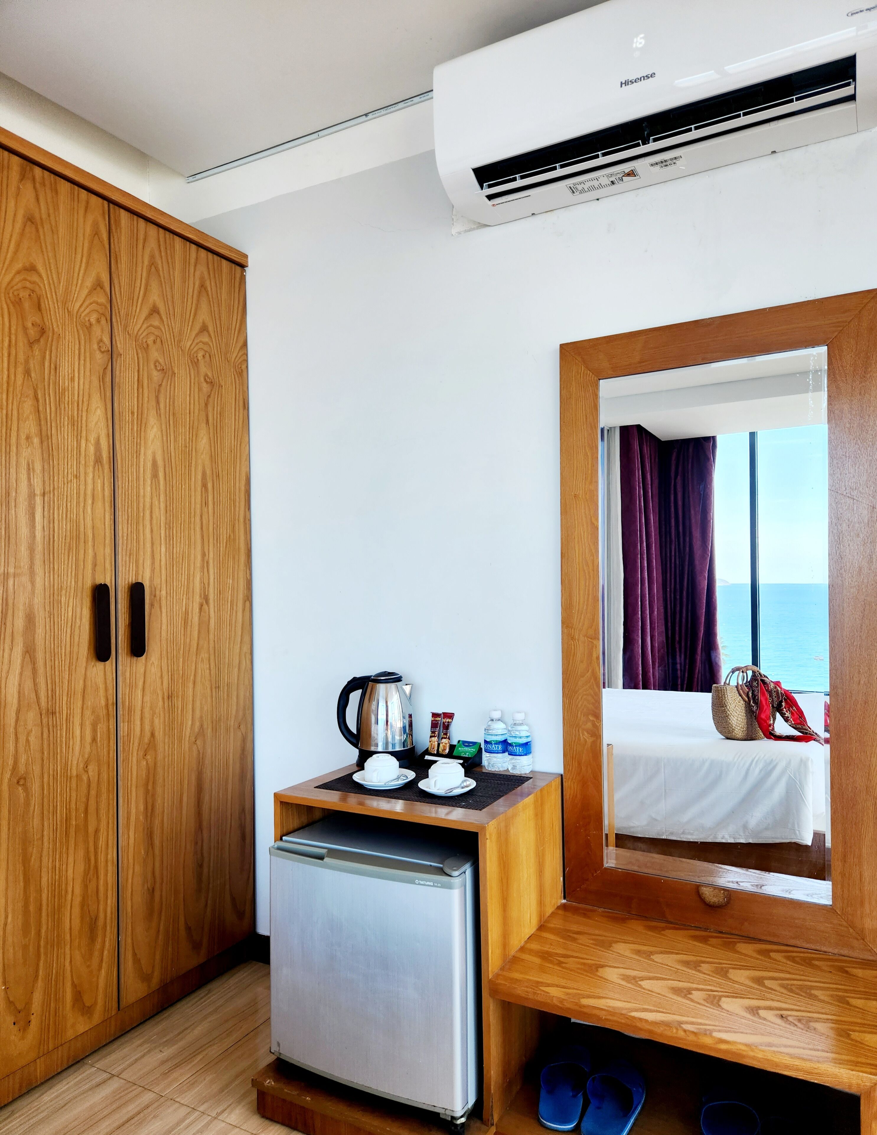 A compact hotel room with a wooden wardrobe, a small kitchenette, a bed with a view of the sea, and a window with purple curtains.