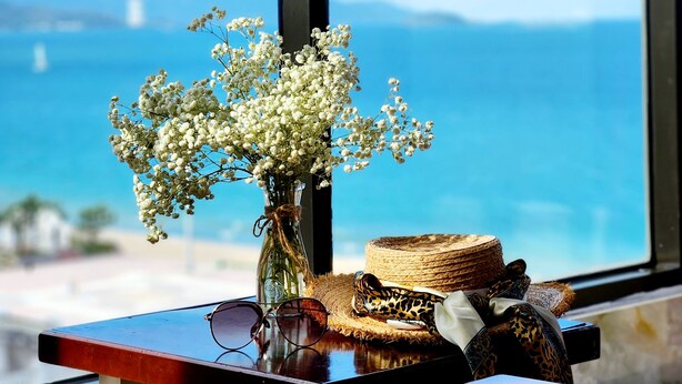 A table with a vase of white flowers, a pair of glasses, and a woven basket with a leopard print bag and a white cloth. - Happy Light Central Nha Trang (Nha Trang)