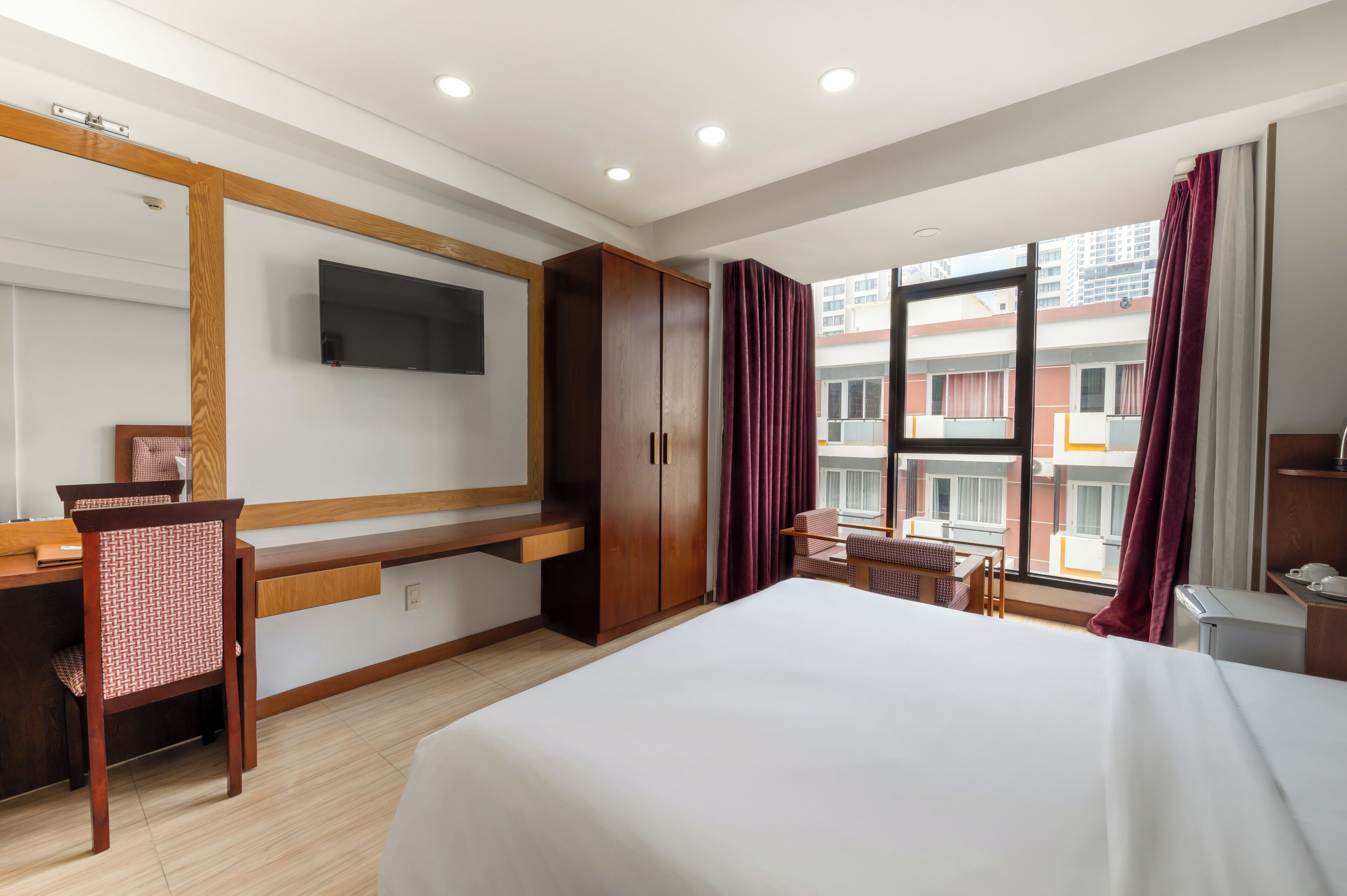 A hotel room with a bed, a desk with a chair, a wardrobe, a TV mounted on the wall, and a window with curtains.