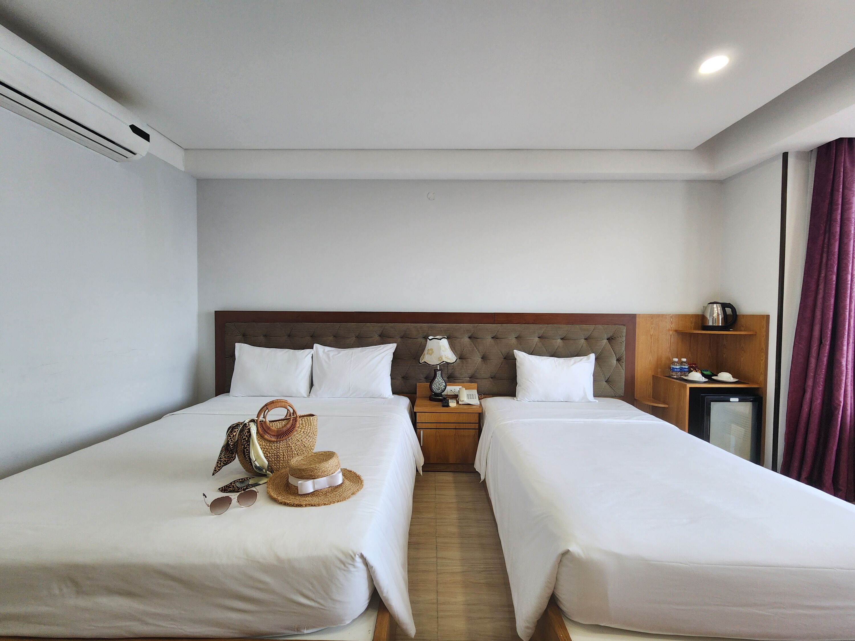 A hotel room with two single beds, a wooden headboard, a bedside table with a lamp, a small TV, and a minibar.