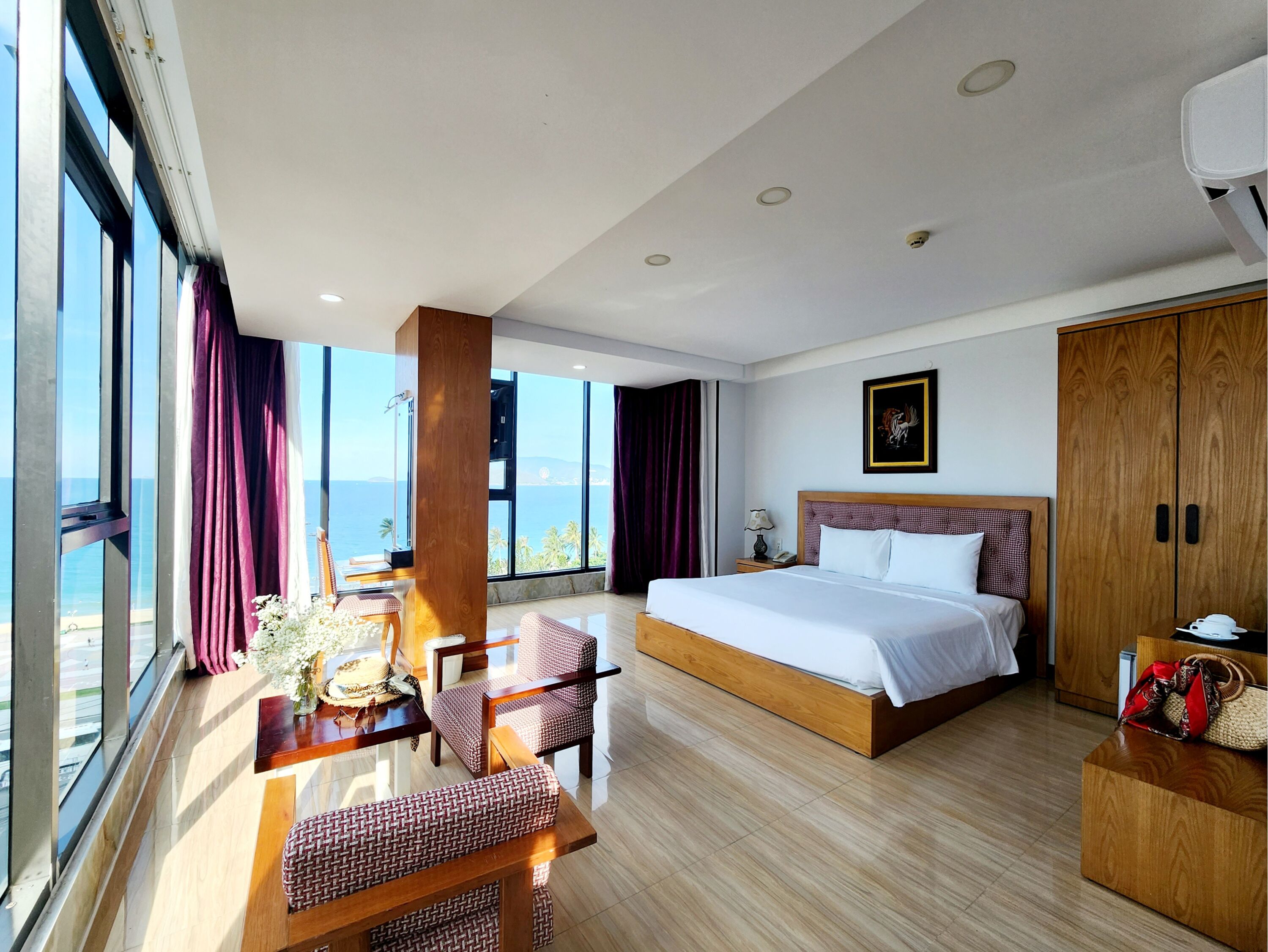 Una habitación de hotel moderna con una cama grande, un armario de madera, un escritorio con una silla y vistas al mar a través de amplios ventanales.